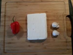 Foto af en Habanero-chili, en blok græsk feta og to fed hvidløg på et skærebræt